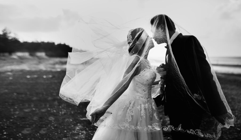 Mariés s'embrassant sur une plage, voile au vent, photographie en noir et blanc